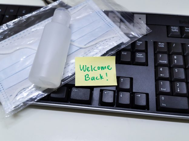 Key board with sticky on it saying 'welcome back' and face masks and hand sanitizer close by